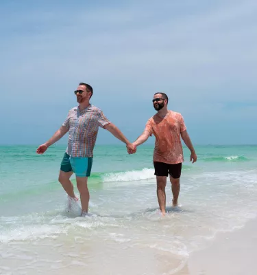 Two men hold hands while walking in shallow water on a pristine beach