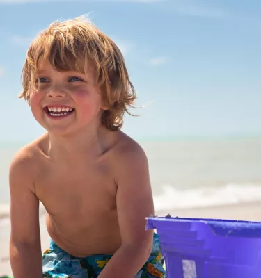 Smiling boy with a purple pail on the beach.