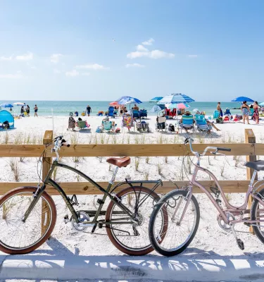 two bikes lean up against a wooden fence near a white-sand beach with umbrellas and people in chairs