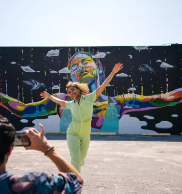 a person taking a photo of a woman wearing a lime green jumpsuit with her arms outstretched in front of a mural of a black woman with her arms outstretched