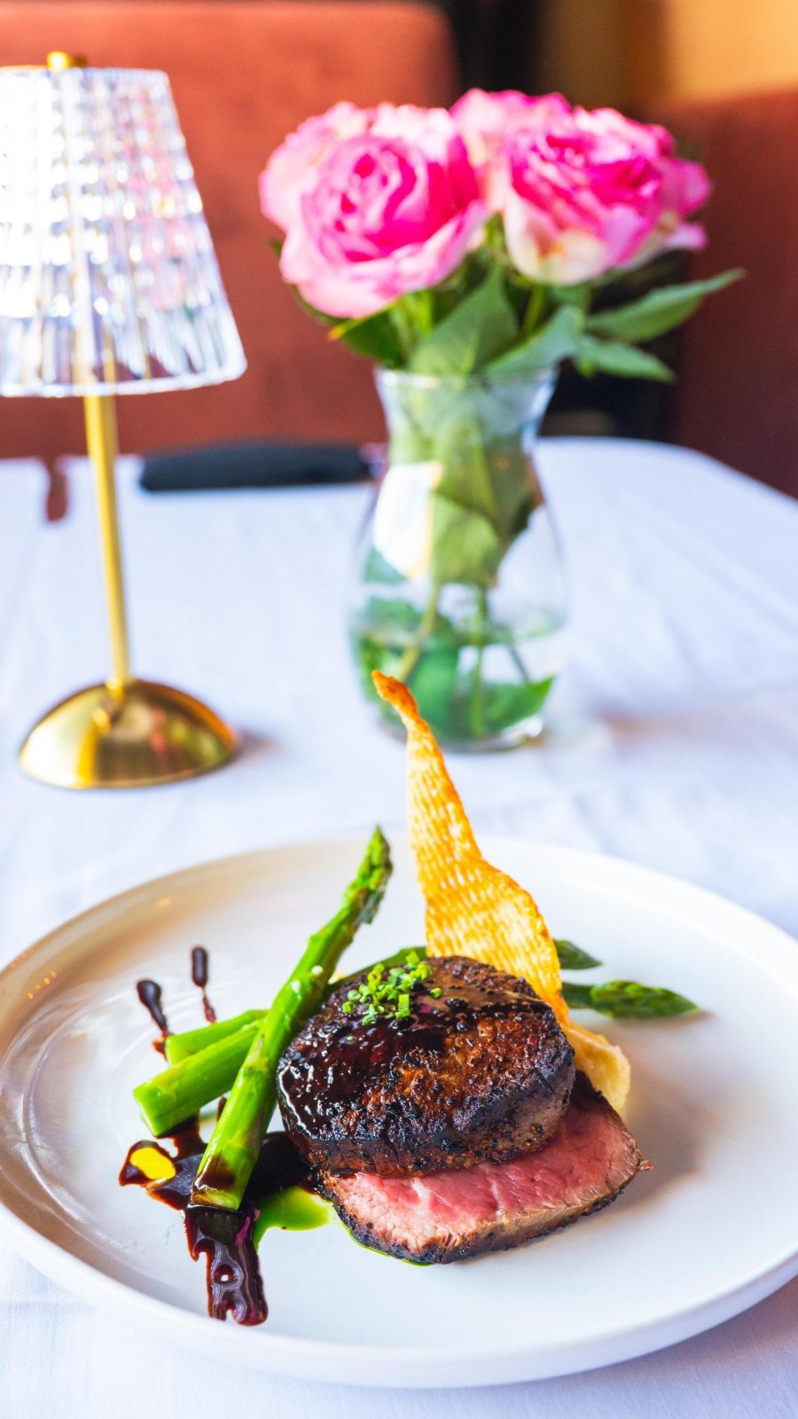 White tablecloth with plate of food on top with steak and asparagus leaning on meat. Table has small lamp and vase with bright pink roses in background.