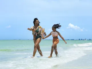 a happy mom and daughter hold hands and smile while walking at the water's edge at the beach