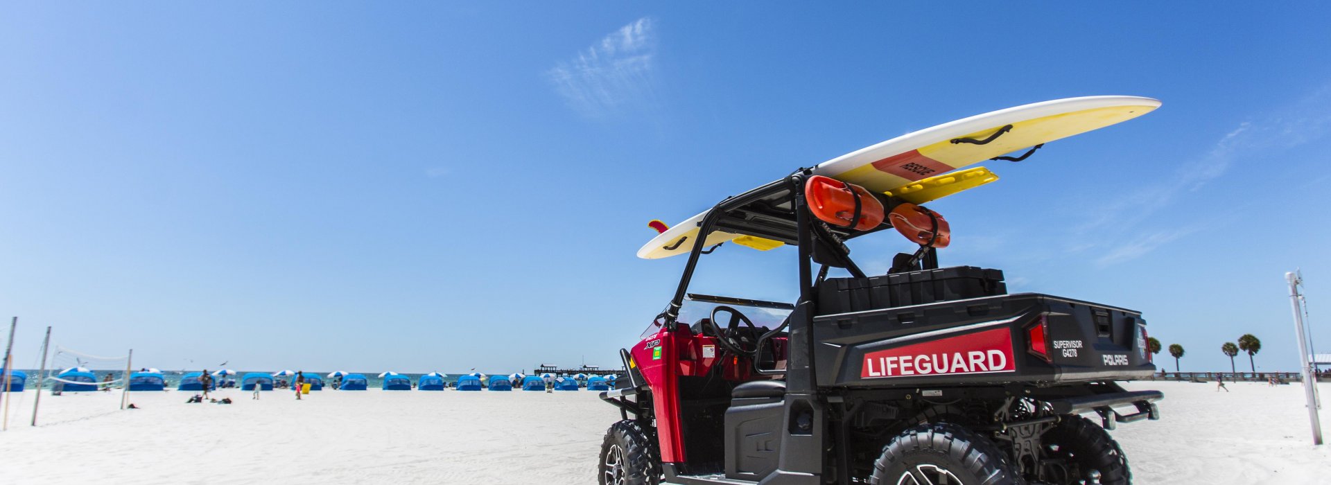 Um buggy salva-vidas estacionado na praia com uma prancha de surf em cima. Cabanas podem ser vistas à distância.