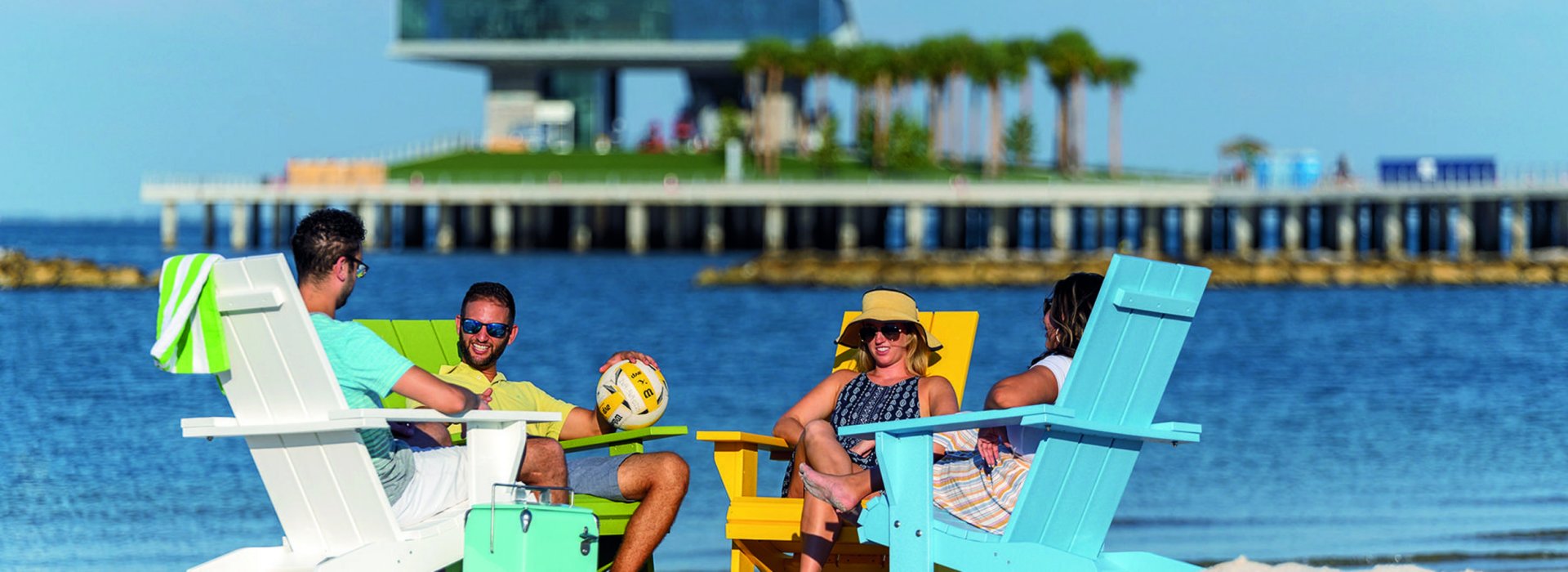 Two men and two women relax in colorful adirondack chairs on Spa Beach with the bay and the St. Pete Pier behind them