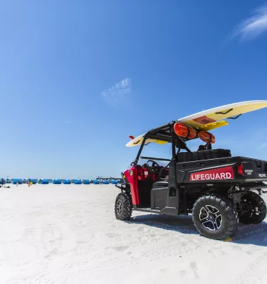 A lifeguard buggy parked on the beach with a surfboard on top. Cabanas can be seen at a distance.