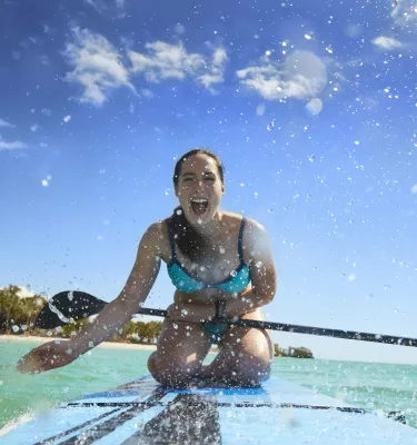 A girl splashing water at the camera while kneeling down on a stand up paddleboard.
