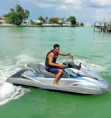 A young man riding a jet ski. Houses and a pier can be seen at a distance.
