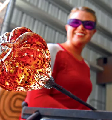 A female glass artist shows off a molten, orange glass art piece in progress at the Morean Arts Center's hot shop in St. Pete