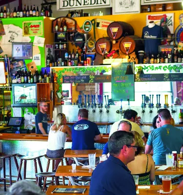 Innenansicht einer Brauereibar mit Menschen, die an der Bar und an den Tischen sitzen. Rückwand gefüllt mit Bärenflaschen, Bierutensilien, Postern, Instrumenten und mehr