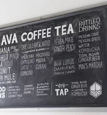 Bild einer schwarzen Kreidetafel an einer Wand mit der Aufschrift KAVA-KAFFEE-TEE in Fettschrift und einer Speisekarte, die den Rest der Tafel vor einer weißen Wand ausfüllt