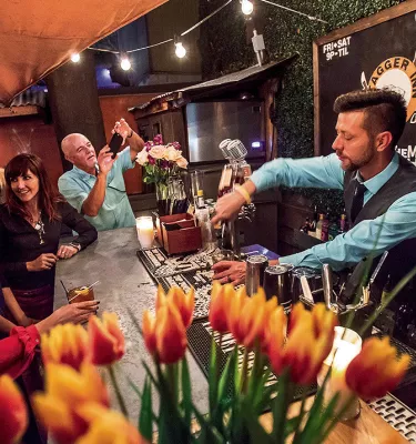 Eine Gruppe von Menschen sitzt an einer Bar, während ein Barkeeper Getränke einschenkt. Im Vordergrund steht eine Vase voller Lilien