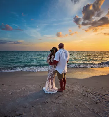 Bride and groom hugging each other and watching the sunset at the beach.