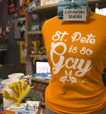 A close view of an orange t-shirt that says "St. Pete is so Gay". Other merchandise and souvenirs are at display.