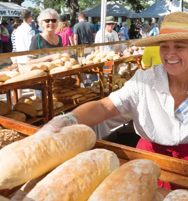 A lady wearing a hat, selling bread with a big smile. Many types of breads are on display.