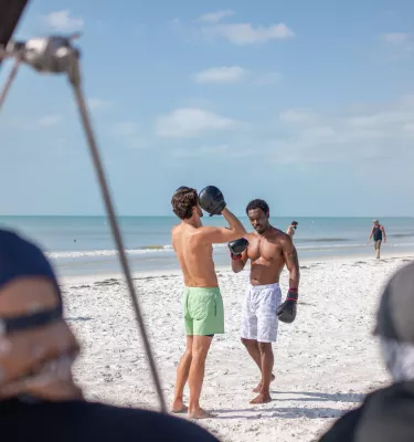 Two men box on the beach in the background with video equipment in the foreground.