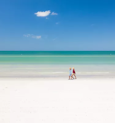 Two beachgoers walk along the white sandy shoreline and emerald green waters of St. Pete Beach on a bright sunny day.