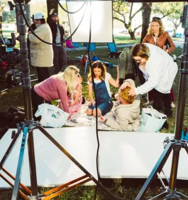 A family sits on a blanket on the grass outside while people setup cameras, hair, and makeup.