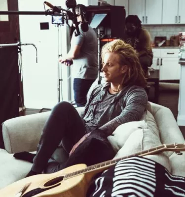The singer sits on a couch next to a guitar with a filming crew in the background.
