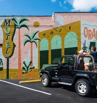 A man and woman in a black jeep park in front of a vintage-themed mural with signs for "motel," "oranges" and "all are welcome here"