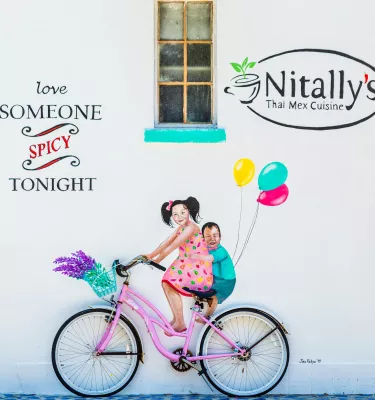 Colorful painting on a white wall of two kids riding a pink bike with balloons on the back