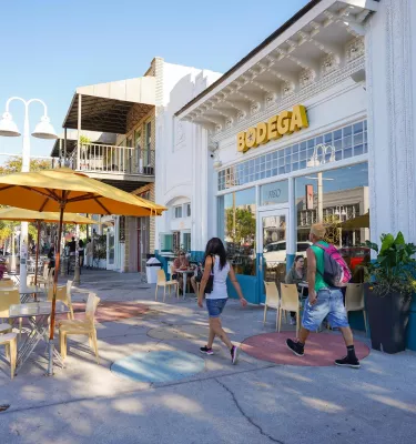 Two people in casual summer clothes walk past bodega in the EDGE District of St. Pete. Tables with yellow umbrellas are on the sidewalk