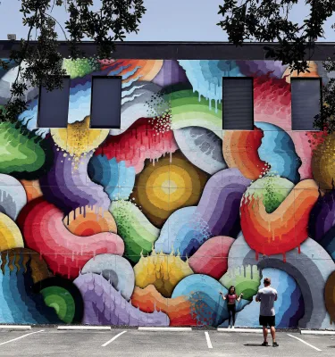 a man takes a photo of a woman in front of an enormous and colorful mural named "Eye of the Storm" in St. Pete's Central Arts District
