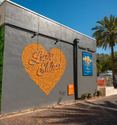 a mural of an orange heart that says Love Shines on a dark gray building with a blue sign and a palm tree to the right