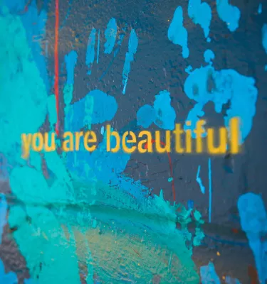 a portion of a large mural displays the words "you are beautiful' in yellow lowercase letters against a background of painted blue, green and red handprints on a dark background