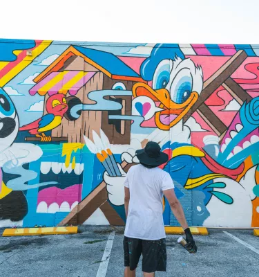 an artist with a spray can of paint in his right hand stands back to look at his work of a cartoonish mural in the EDGE District St. Pete