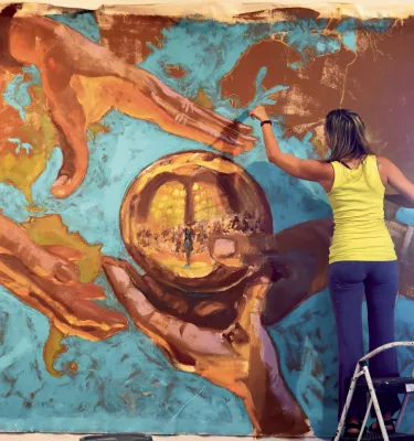 artist Carrie Jadus in a yellow shirt and bluejeans standing on a ladder painting a mural in st. pete