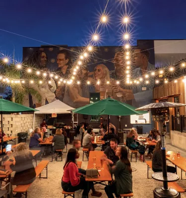people drinking beer at long picnic tables outside with twinkling lights and a mural in the background at Grand Central Brewhouse