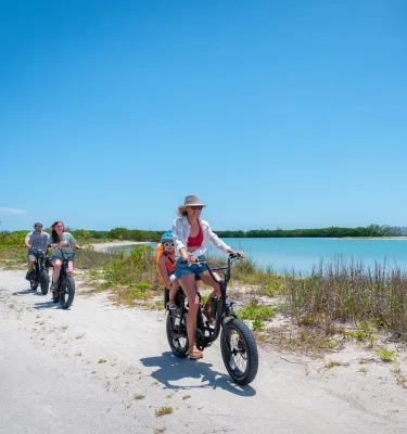 A woman and a baby on an e-bike ride next to blue-green water and sea oats with other family members biking behind them at Fort De Soto Park