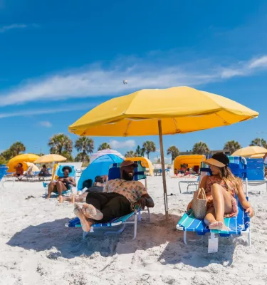 a couple relaxes on lounge chairs under a bright yellow umbrella on the white sands of Madeira Beach