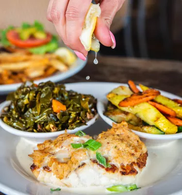 a woman squeezing a lemon on top of a plate of grilled fish, sauteed greens and mixed veg at Hog Island Fish Camp in Dunedin