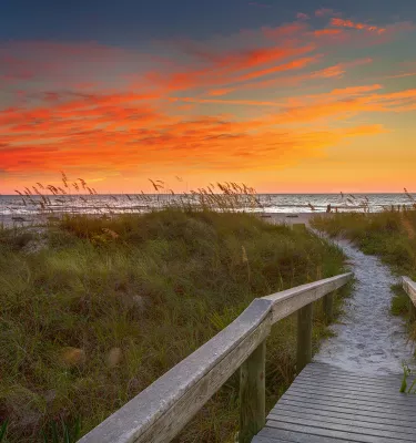 a wooden boardwalk through sea oats and other vegetation leads to the Gulf, the boardwalk is sandy, the sky is just after sunset with orange and blue colors