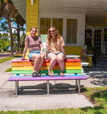 two women in shorts and tshirts sit on top of a bench painted in rainbow colors in front of a bright yellow inn next to a street