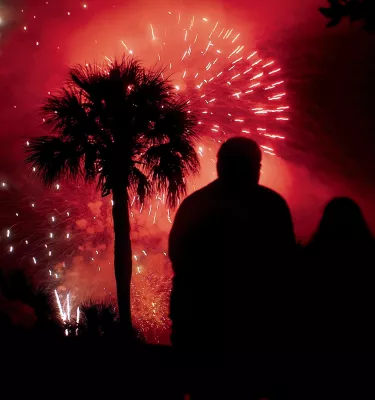 Fireworks explode in red sky with silhouette of palm tree and two people