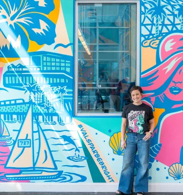 a person in jeans and a black t-shirt stands in front of a pastel colored mural named "be bright" at St. Petersburg Clearwater International Airport
