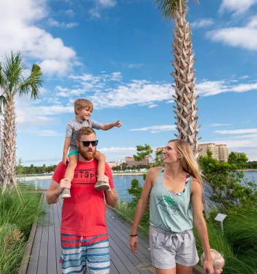 father with little boy on his shoulders walking next to a woman on a boardwalk surrounded by palm trees near the water