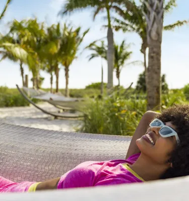 Woman with sunglasses laying in a hammock with the beach and palm trees in the background
