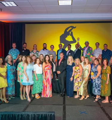 a group of well dressed people standing together for a photo in front of a large screen with an image of a dancer at sunset
