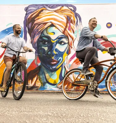 two men riding bikes in front of a mural of an African woman with a turban