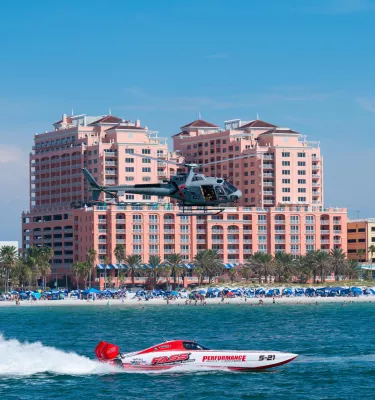 a race boat at top speed in the Gulf of Mexico with a helicopter flying overhead and the beach and hotels in the background
