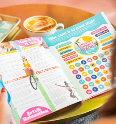 a woman reading a magazine with brightly colored pages at a small table set with a latte and a buttery croissant