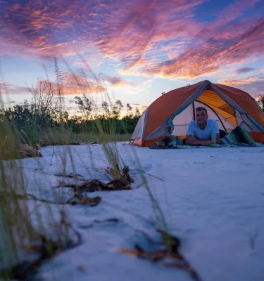 a young man relaxes inside his tent on the beach at Shell key with a dramatic pink and blue sky behind him