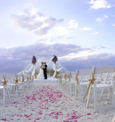 a couple embraces after being married on the beach at Honeymoon Island at sunset, with white chairs and pink rose petals as decorations