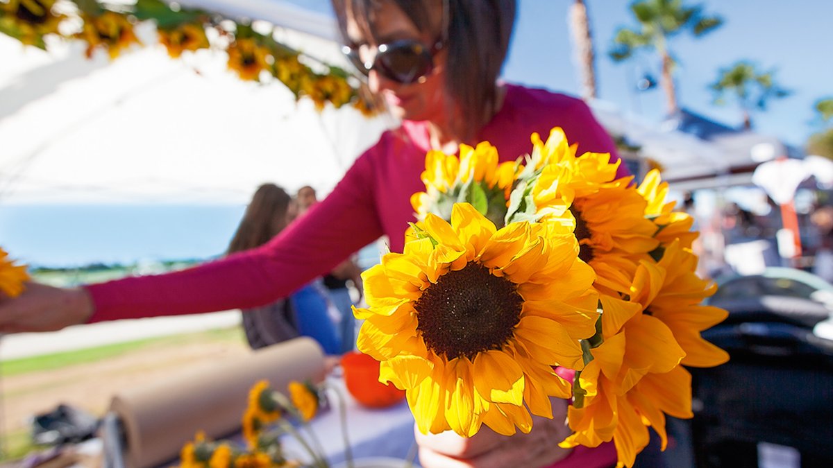 A woman holds a bundle of sunflowers at a sunny, open-air market.