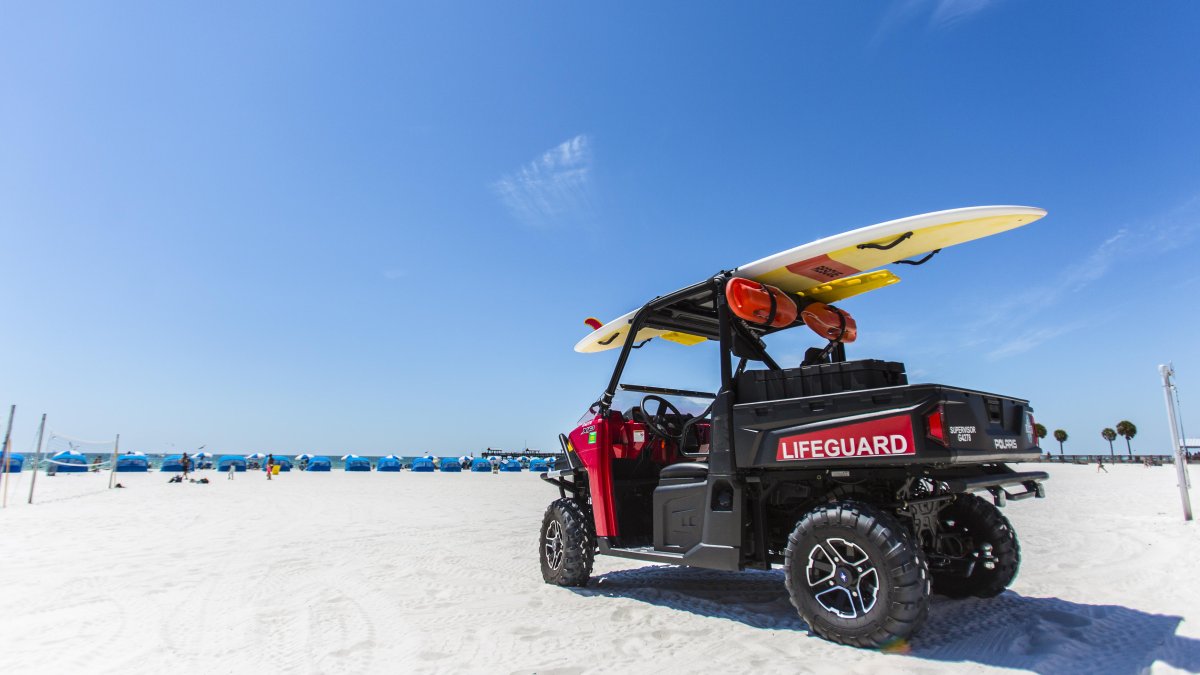 A lifeguard buggy parked on the beach with a surfboard on top. Cabanas can be seen at a distance.