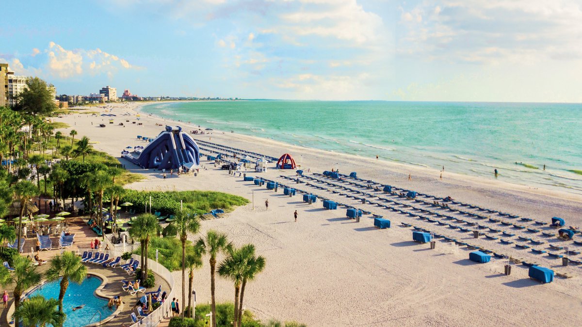 Overview of Tradewinds Resort beachfront pool showing blue cabanas, a water slide and aqua-marine water.