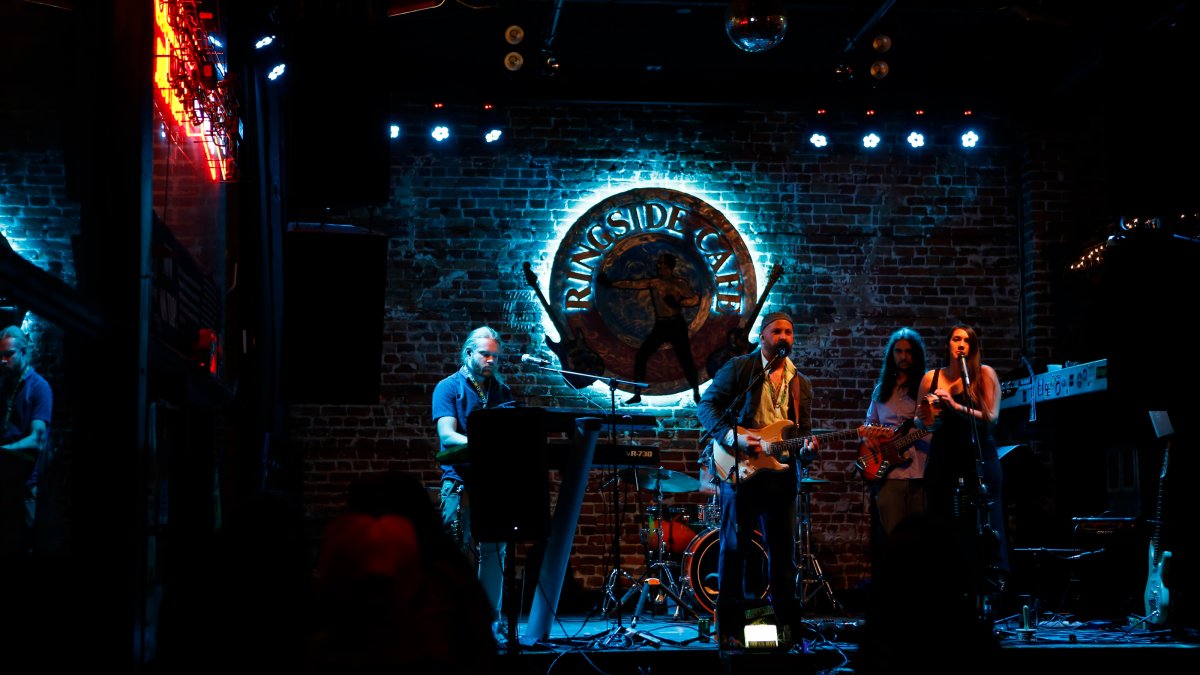 A band playing at night at the Ringside Cafe. A blue sign with the logo is shining behind the musicians.